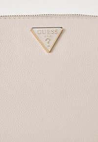 Světle béžová texturovaná kožená taška s zlatou trojúhelníkovou logovou deskou s nápisem "GUESS" a "?". Zipové uzavření podél horního okraje.