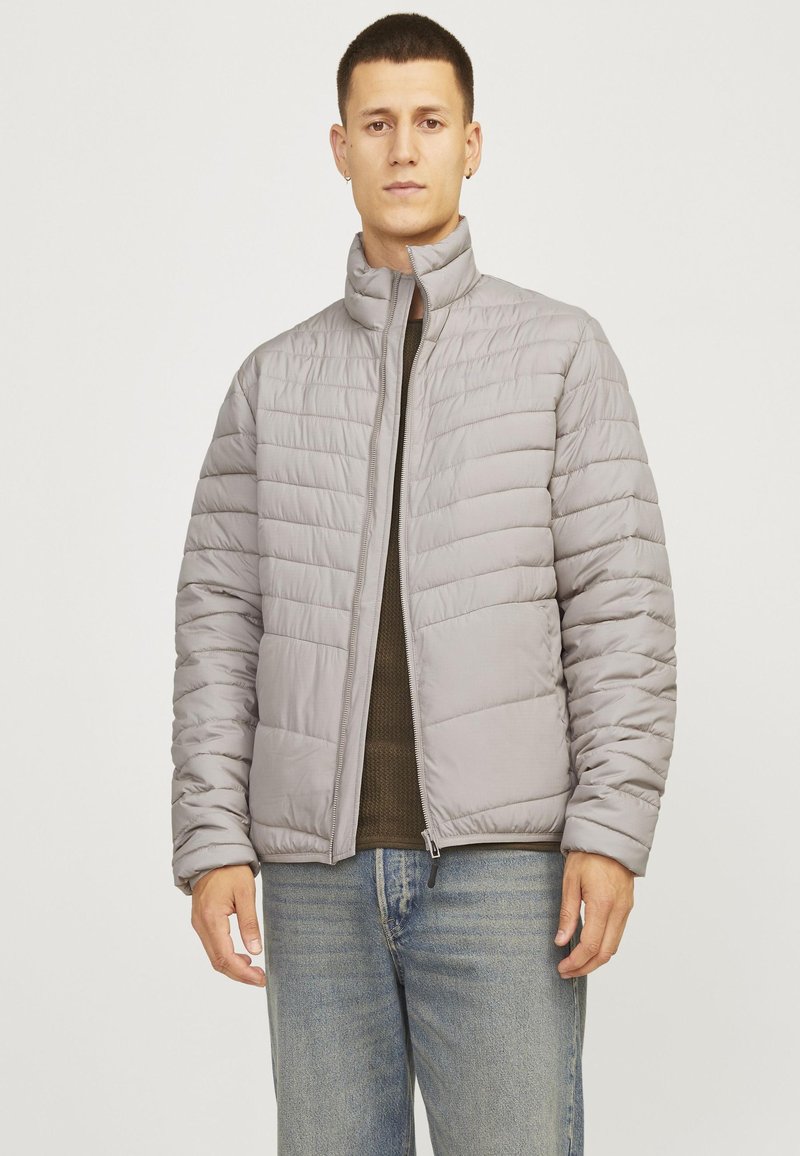 Light gray puffer jacket with a high collar, horizontal quilted pattern, and front zipper. Worn over a dark green top and denim jeans.