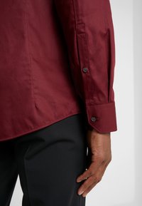 Camisa burdeos de manga larga con una textura suave, puños de un solo botón y dobladillo curvado, combinada con pantalones negros.