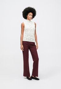 Kvinne med stor afro som har på seg en ærmeløs hvit blonde topp, maroon vide bukser og svarte naglede flate sko, som står mot en ensfarget bakgrunn.