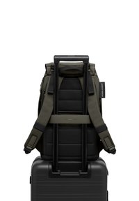 Sac à dos vert foncé avec des straps rembourrés, dos texturé, attaché à une valise rigide noire avec une poignée télescopique.