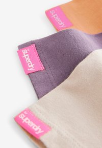 Tre bomullströjor i orange, lila och beige, med framträdande rosa etiketter som har "superdry"-logotyp längs kanterna.