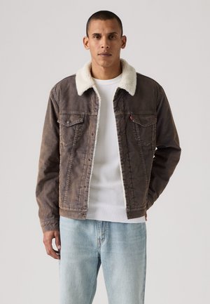 Homme portant une veste en denim marron avec col en polaire blanc par-dessus une chemise blanche texturée et un jean bleu clair sur fond uni.