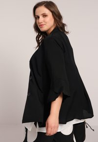 Veste noire, surdimensionnée avec de larges manches, en tissu texturé, présentant deux grands boutons à l'avant, superposée sur une blouse blanche.