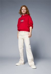Sweatshirt à capuche rouge avec logo, jeans blancs évasés et baskets blanches. Le mannequin a des cheveux bouclés et porte des lunettes, se tenant devant un fond gris.