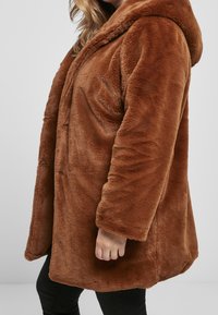 Brun faux fur frakke med hætte, der har en blød tekstur og en afslappet pasform. Inkluderer en frontlukning med spænde og sidelommer.