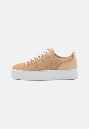 Beżowy niski sneaker o koronkowej fakturze z białymi sznurowadłami i grubą białą gumową podeszwą na jasnoszarym tle.