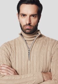 Beżowy sweter w splot kablowy z wysokim kołnierzem i zapięciem na zamek, z teksturowanym wzorem i ściągaczami na rękawach, noszony na beżowym golfie.