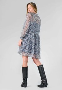 Vestido mini semitransparente con estampado floral en tonos azules y rosas, presenta mangas largas, una cintura fruncida y una falda acampanada, combinado con botas negras hasta la rodilla.
