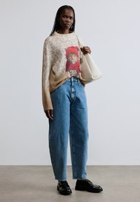 Beež värvitud kampsun leopardiprindiga, millel on värvikas kassi graafika, sobitatud siniste lahtiste teksade ja mustade kingadega, kaasas valge kott.