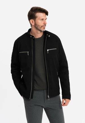Veste noire avec un col montant, des accents de fermeture éclair métalliques et deux poches poitrine zippées. Fabriquée dans un tissu texturé solide.