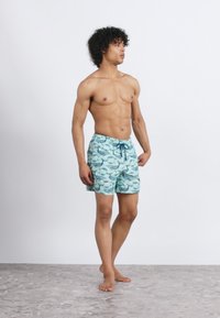 Muž bez košele stojí naboso na kamennej podlahe, oblečený v svetlomodrých plavkách s tmavomodrým vzorom rýb a sťahovacím pásom.