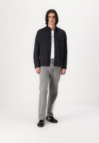 Must must shirts, kus on kaks rinna taskut, koos hallide teksade ja mustade tossudega, millel on valged triibud. Mudel seisab valge tausta ees.