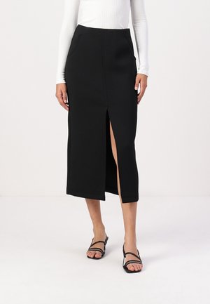 Kobieta ubrana w czarną spódnicę midi z rozcięciem z przodu, w połączeniu z białą bluzką z długim rękawem i czarnymi sandałkami na obcasie z paskami.