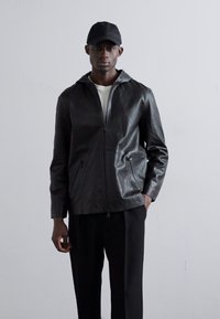 Jaqueta de couro preta com capuz, apresentando um fecho frontal e dois bolsos laterais. Usada sobre uma camisa branca e combinada com calças pretas.