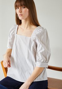Blusa às riscas com decote quadrado, mangas bufantes e um tecido leve. As riscas são azuis sobre um fundo claro.