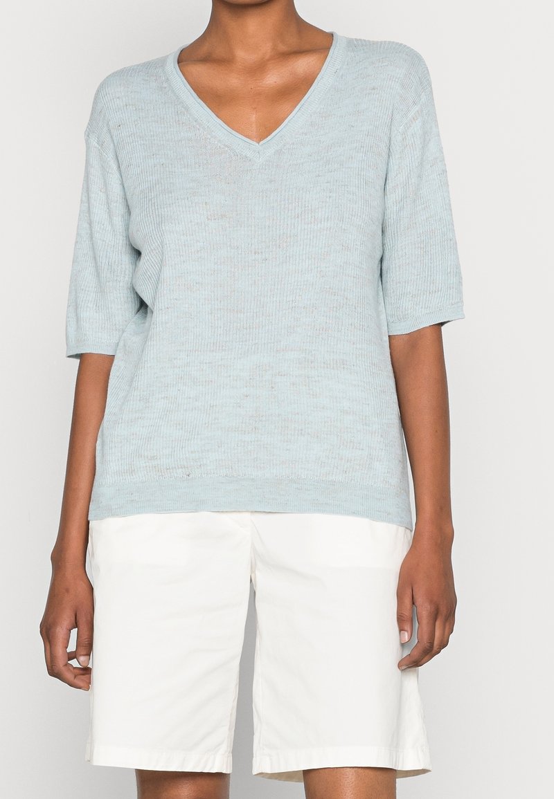 Personne portant un pull à encolure en V côtelé bleu clair avec des manches mi-coudes et un short blanc d'une longueur aux genoux, debout devant un fond uni.