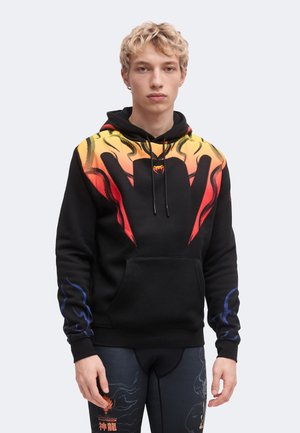 Joven con una sudadera negra con capucha con diseños de llamas amarillas y rojas en el pecho y patrones de llamas azules en las mangas.
