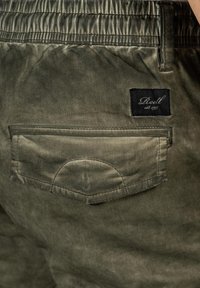 Olivgrüne Shorts mit strukturierter Oberfläche, ausgestattet mit einer Gesäßtasche und einem schwarzen Label mit weißer Schrift. Elastischer Bund.