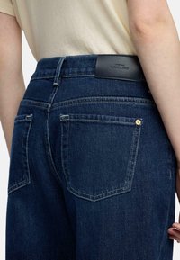 Ciemnoniebieskie dżinsy z denimu o prostych nogawkach, tylne kieszenie z detalami przeszyć oraz czarną skórzaną etykietą na pasku w talii.