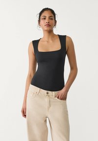 Černý bodysuit s obdélníkovým výstřihem vyrobený z hladké, strečové tkaniny, v kombinaci s světlými béžovými kalhotami s vysokým pasem a širokými nohavicemi.