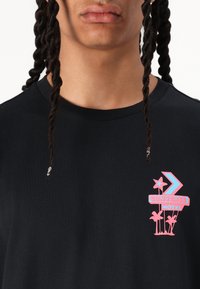 Sort bomulds T-shirt med et farverigt grafik med en stjerne og palmetræer. Designet inkluderer teksten "CONVERSE MOTEL" i pink og blå accenter.
