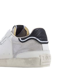 Bílé tenisky s semišovými a texturovanými černými hadími akcenty na patě. Obsahují polstrovaný límec a texturovanou podrážku s embosovaným nápisem "1973".