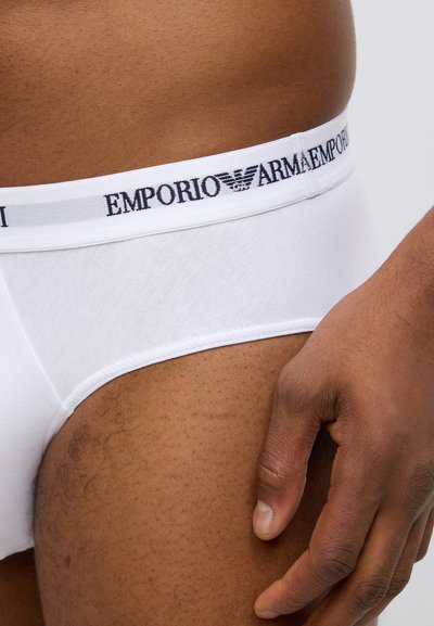 Caleçon blanc en coton avec une ceinture noire arborant le logo Emporio Armani. Texture lisse avec une coupe ajustée. Peau visible au niveau de la cuisse.