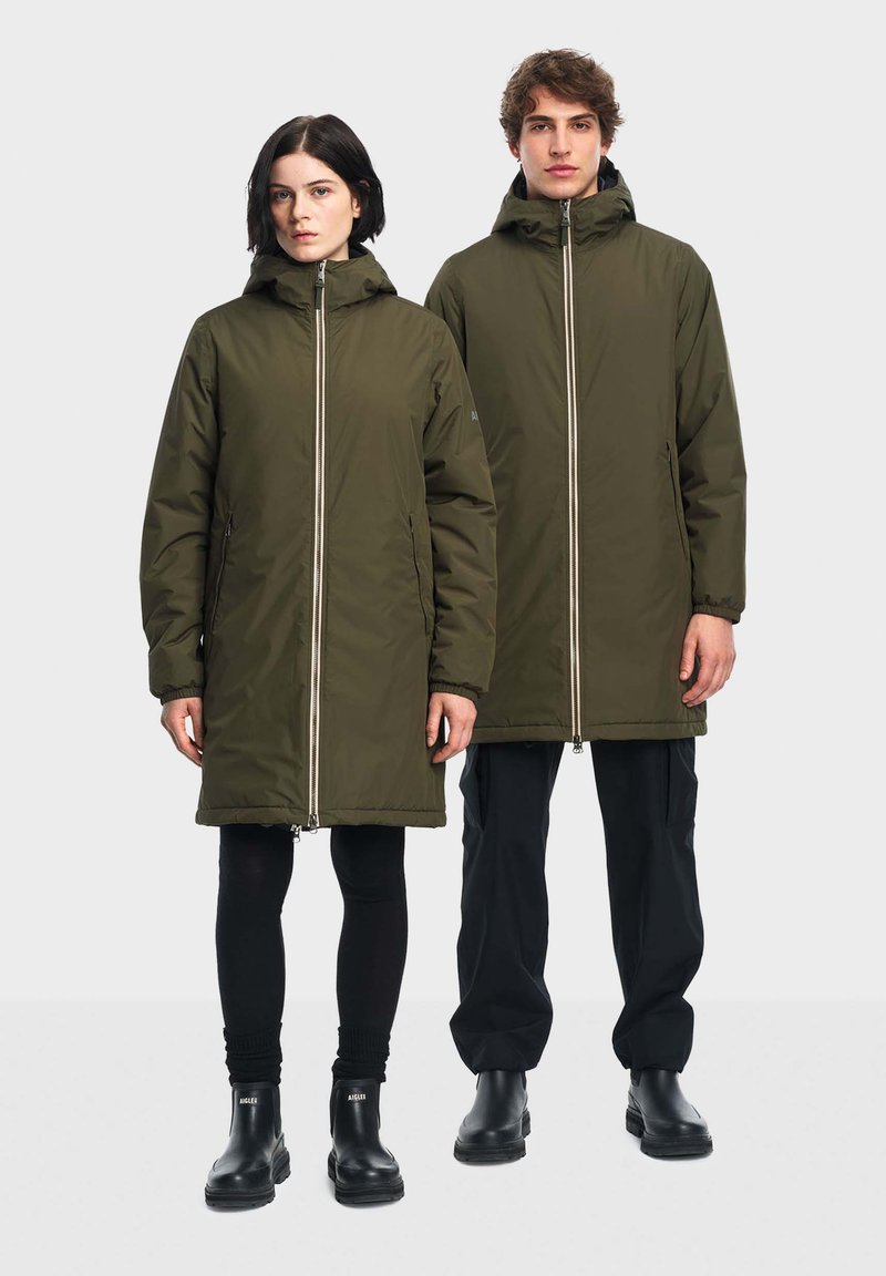 Parka vert olive zippée avec capuche, coupe ample et poches latérales. Porté sur des legging noirs et des bottes noires épaisses, avec une texture lisse.