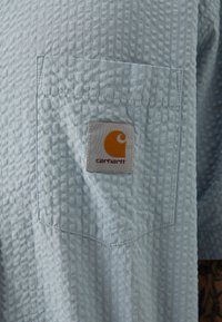 Chemise à manches courtes bleu clair, en tissu texturé avec un motif à carreaux, dotée d'une poche poitrine et d'un patch logo Carhartt en orange.