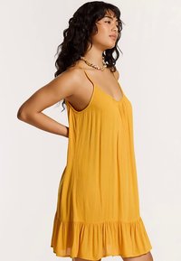 Dzeltens bezroku kleita ar izplestu maliņu, izgatavota no viegla auduma, ar teksturētu virsmu un vienkāršu V-veida kakla izgriezumu.