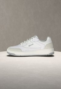 Λευκό sneaker χαμηλού πάτου με γκρι σουέντ λεπτομέρειες, κορδόνια και λογότυπο "Just Cavalli" στην πλευρά, τοποθετημένο σε ουδέτερο γκρι υπόβαθρο και επιφάνεια.