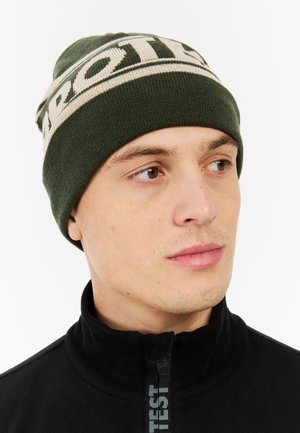 Green knitted beanie with cream logo band, tight fit, ribbed texture. Worn with a black zip-up fleece top. Close-up of face.