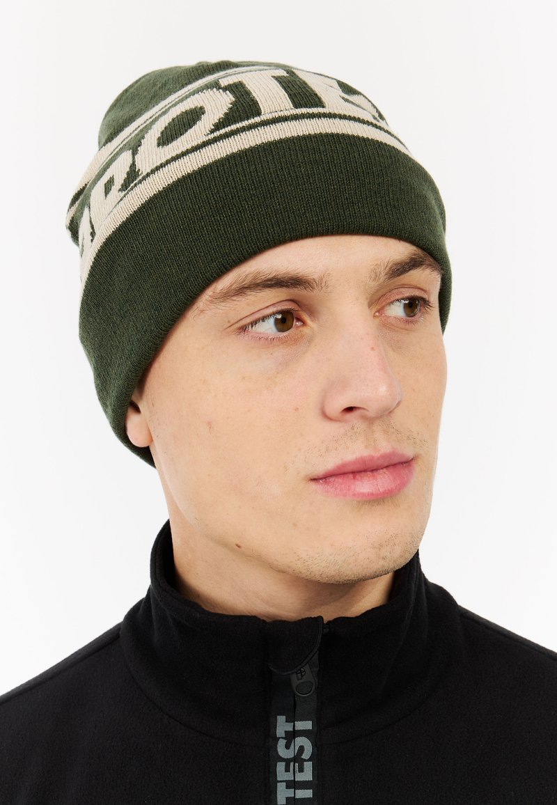 Zelena pletena kapa s kremastim logom, uskog kroja, rebraste teksture. Nosena uz crni zip-up flis duksericu. Krupan plan lica.