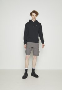 Schwarzer Hoodie mit einer Vorderentasche, graue Cargo-Shorts und schwarze Knöchelstiefel. Das Model steht vor einem schlichten weißen Hintergrund.
