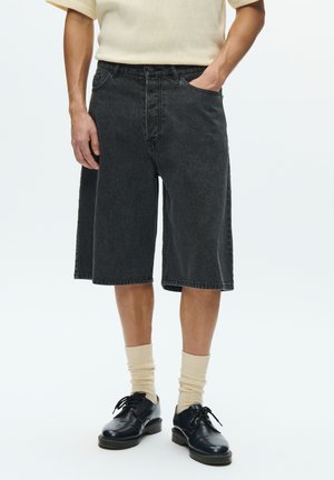 Shorts en jean noir, longueur au genou avec une coupe ample, comprenant cinq poches et une fermeture à bouton. Portés avec des chaussettes claires et des chaussures noires.