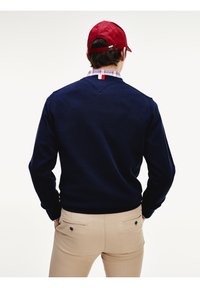 Man som bär en marinblå tröja, beige byxor, röd keps och rutmönstrad skjorta, står med händerna i fickorna och vänder ryggen mot kameran framför en vit bakgrund.