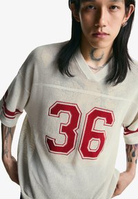 T-shirt en jersey crème en maille avec des accents rouges, présentant le numéro 36 de façon proéminente sur le devant. Manches courtes et design en v.