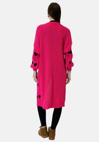 Lange roze gebreide cardigan met zwarte hartpatronen op de mouwen, geribbelde boorden en een losse pasvorm. Draagt over zwarte leggings en bruine laarzen.