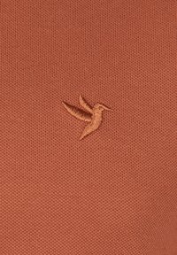 Tissu tricoté couleur rouille présentant un logo de colibri brodé en détail dans une teinte plus claire, positionné sur le côté gauche.