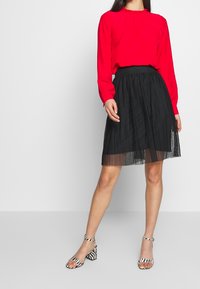 Rote Bluse mit langen Ärmeln und gerafftem Ausschnitt, kombiniert mit einem schwarzen, plissierten Rock. Das Modell trägt gestreifte Sandalen mit Blockabsatz.