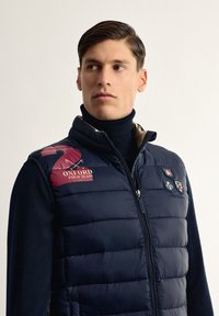 Chaleco acolchado azul marino con un alto cuello, logotipo rojo "Oxford Polo Team" y múltiples parches bordados en el pecho, con cierre de cremallera.