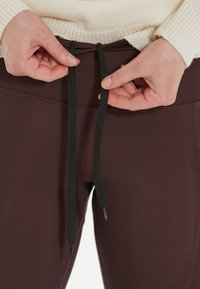Leggings de sport bordeaux en tissu lisse et extensible ; des cordons noirs sont noués à la taille, avec des détails de couture subtils.