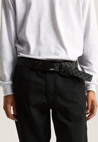 Black checkered belt with a rectangular buckle, featuring a Vans logo. Worn over black pants, paired with a light gray long-sleeve shirt.