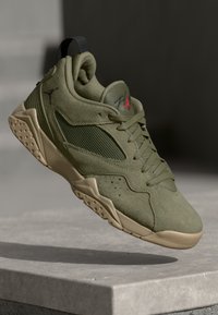 Sneaker de ante verde oliva y malla con suela marrón claro y logo Jumpman rojo, flotando sobre una superficie de piedra con sombra abajo.