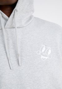 Hele halli kapuutsiga, millel on pehme tekstuur, ja rinnal on valge tikitatud näo logo. Jooned on hallid ja ümarad.