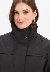 Schwarze Jacke mit hohem Kragen, mit Druckknöpfen, glatter Textur und horizontalen Steppnähten auf der Brust.