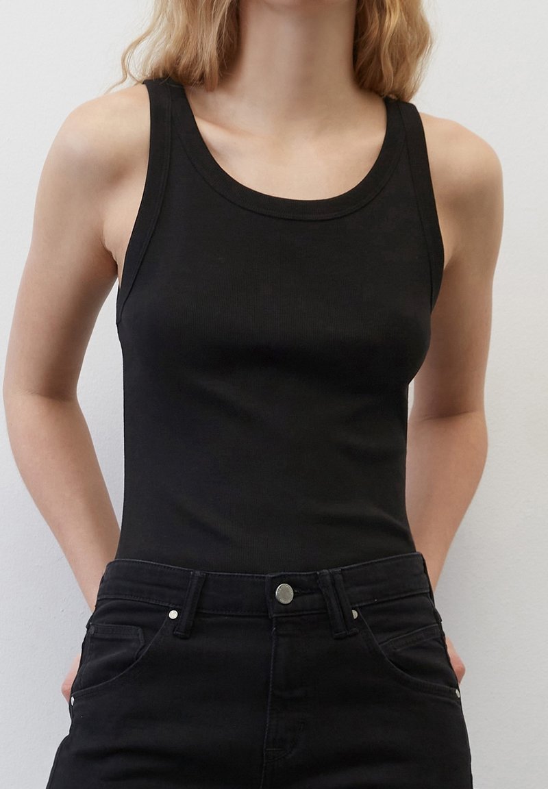 Femme portant un débardeur noir sans manches rentré dans un jean noir taille haute, debout devant un fond blanc uni.