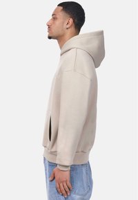 Beige hoodie with a kangaroo pocket, relaxed fit, and ribbed cuffs. Soft material, featuring front embroidery, and a drawstring hood.