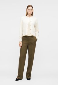 Γυναίκα που στέκεται με τα χέρια στις τσέπες, φορώντας μπεζ ζακέτα με χρυσά κουμπιά, χακί φαρδιά παντελόνια και μαύρα μυτερά παπούτσια σε λευκό φόντο.